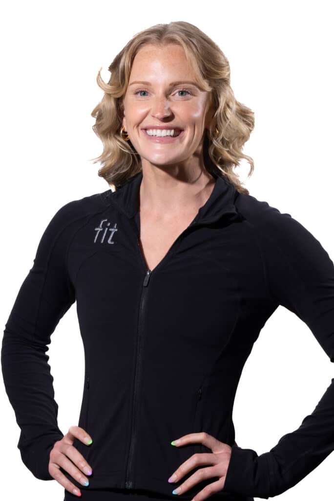 A person with shoulder-length, wavy blonde hair and a black zip-up jacket smiles, standing with hands resting on hips.