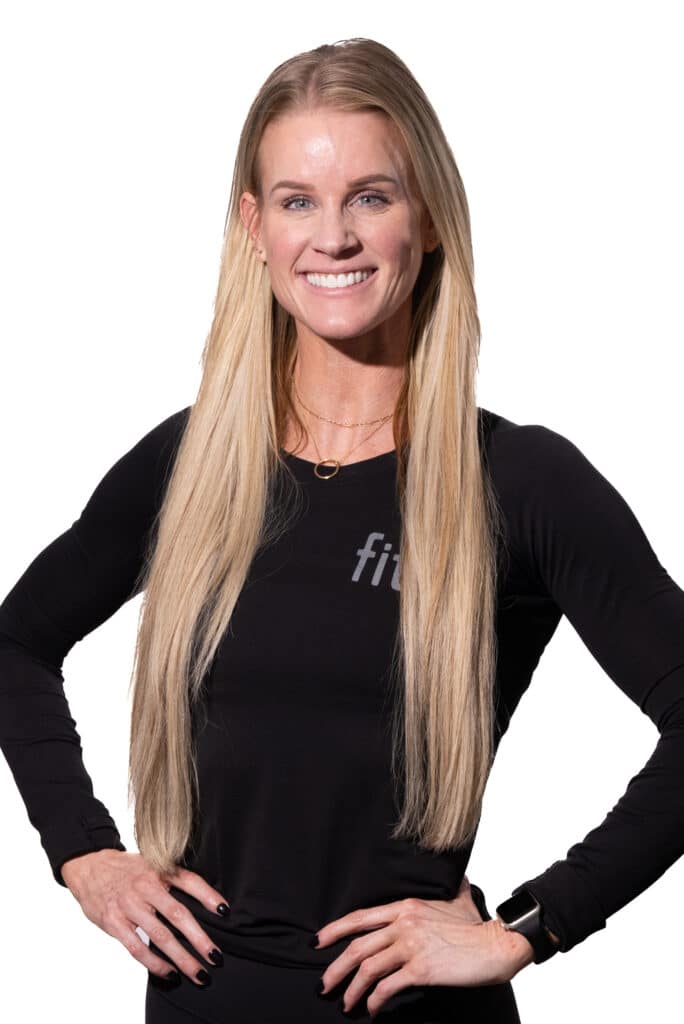 A woman with long blonde hair is smiling, wearing a black long-sleeve shirt with "fit" printed on it and a black wristwatch. She has her hands on her hips.