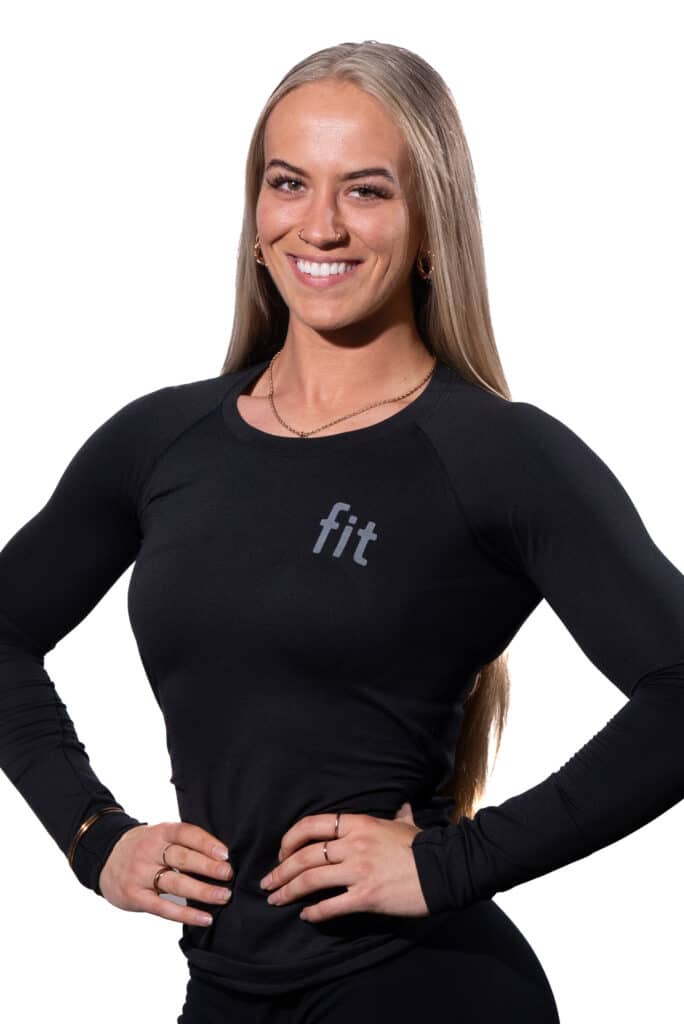 A woman with long blonde hair, wearing a black long-sleeved shirt labeled "fit," is smiling with her hands on her hips.