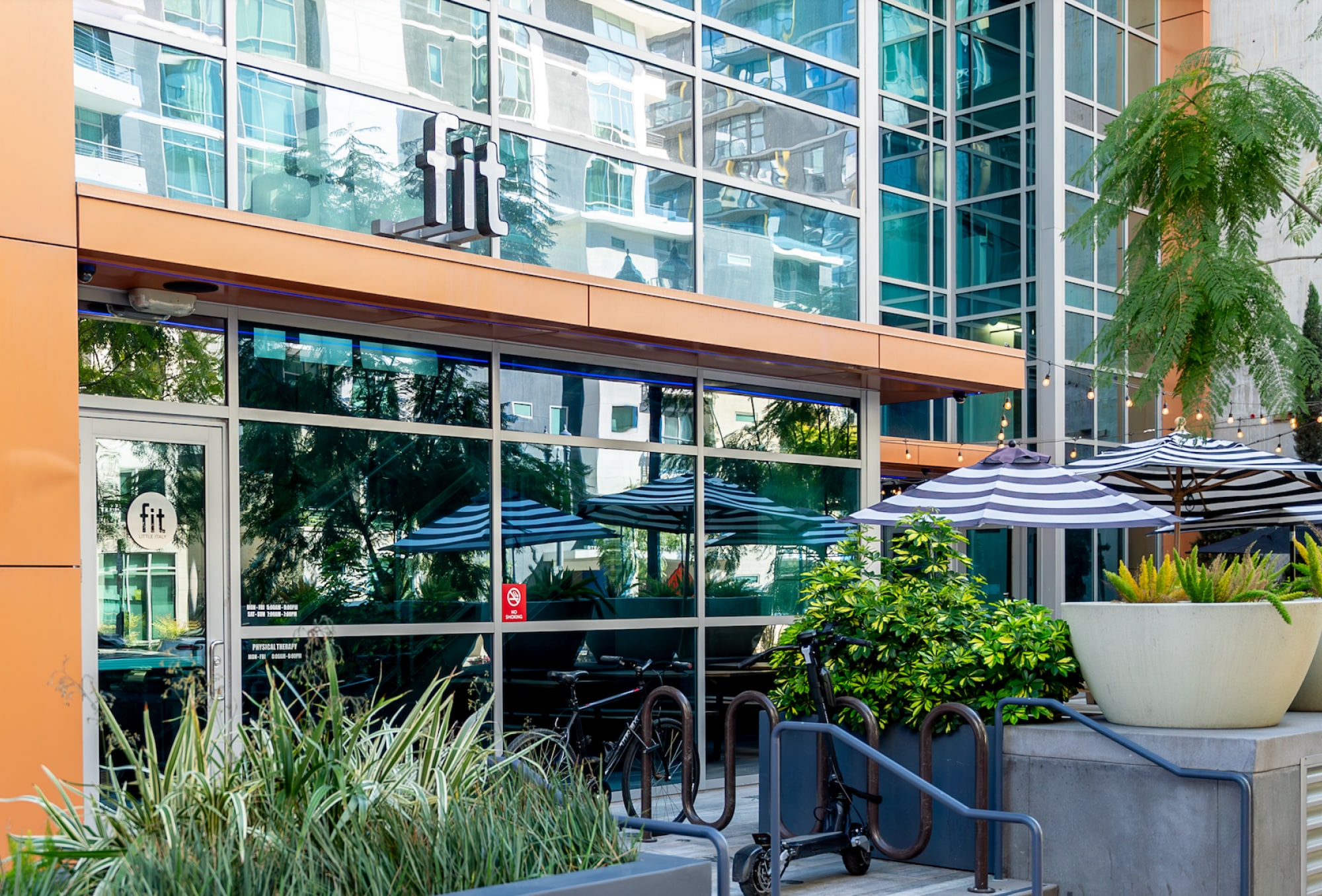 Exterior view of a fitness center with "fit" signage. Features glass windows, outdoor seating with black-and-white striped umbrellas, and surrounding greenery.