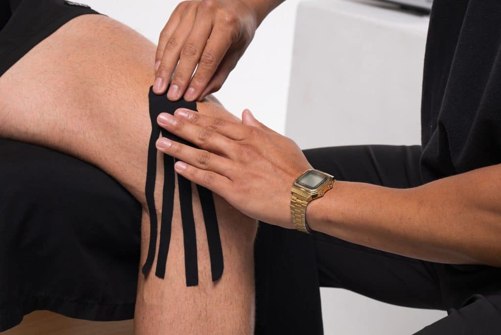 A person wearing a gold watch applies black kinesiology tape to the knee of another individual seated in front of them.