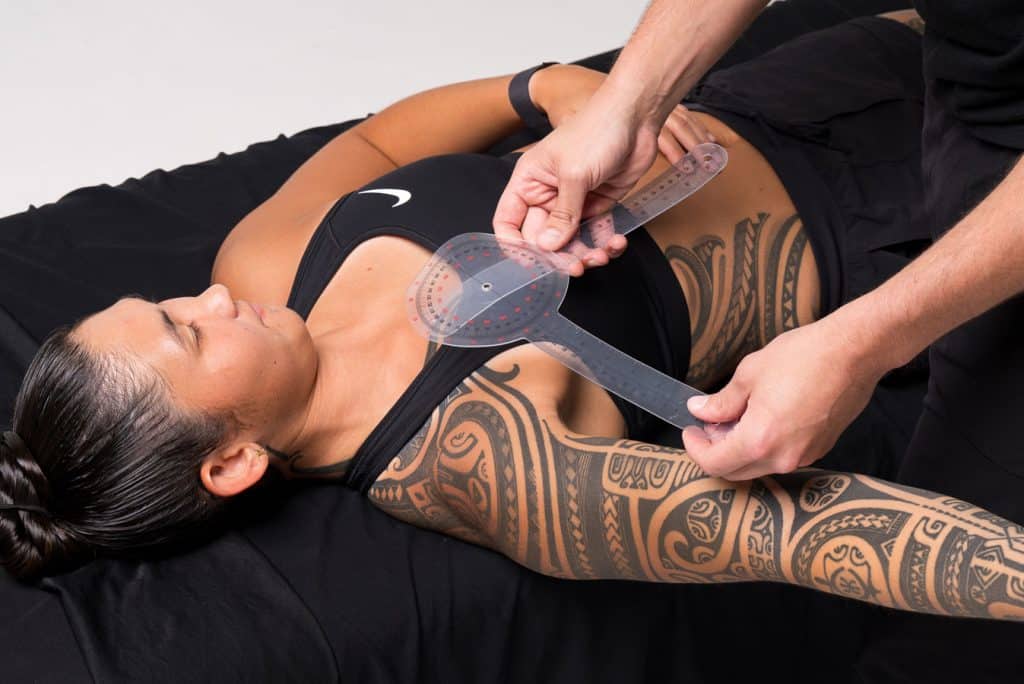 A person with tattoos on their upper body lies on a black surface while another person measures their shoulder angle using a plastic goniometer.