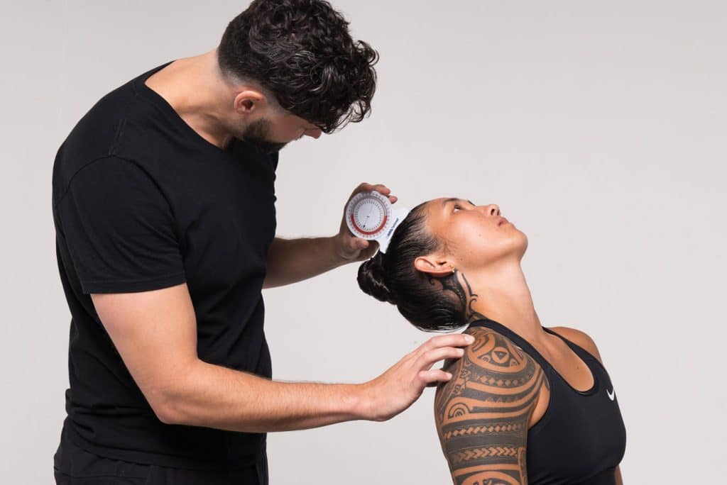 A person in a black shirt measures the neck angle of another person with a protractor. The person being measured has a tattooed arm and is wearing a black tank top.