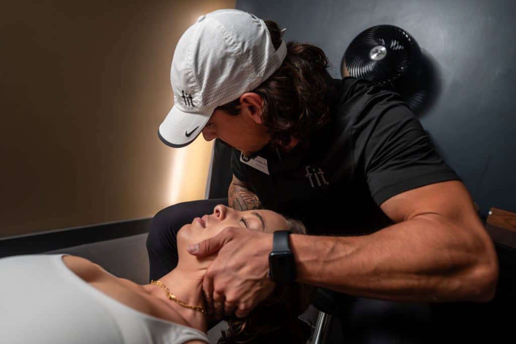 A chiropractor, wearing a white cap and black shirt with "fit" logo, performs a neck adjustment on a female patient lying on a table in a treatment room.