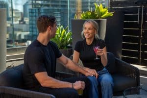 Two people sit together on a rooftop patio. The woman holds a purple drink and smiles, while the man rests his hand on her knee. Green plants and city buildings are in the background.