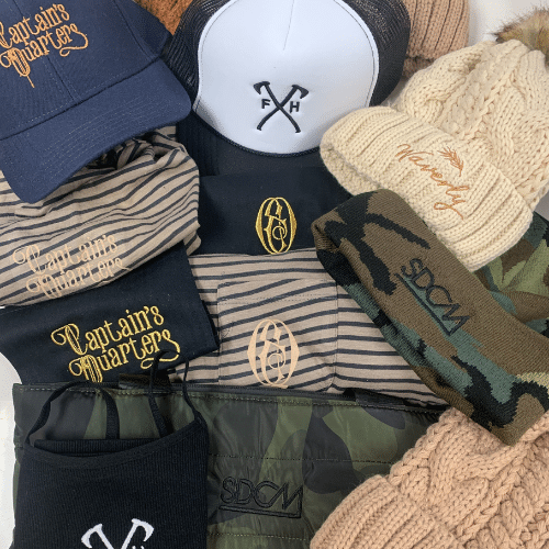A collection of hats, shirts, and headbands with various logos including "Captain's Quarters," "SDCM," and "FH." The items are in navy, beige, black, camo, and knitted patterns.