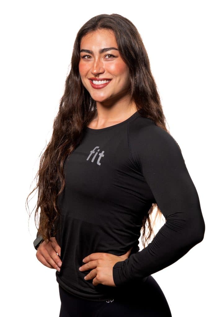 Woman with long hair in black athletic shirt smiling, standing with hands on hips against a white background.